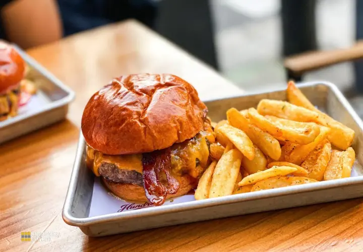 Jugosa hamburguesa con queso fundido y tocino crujiente, acompañada de patatas fritas doradas. Un festín para los sentidos en Hundred Valencia.