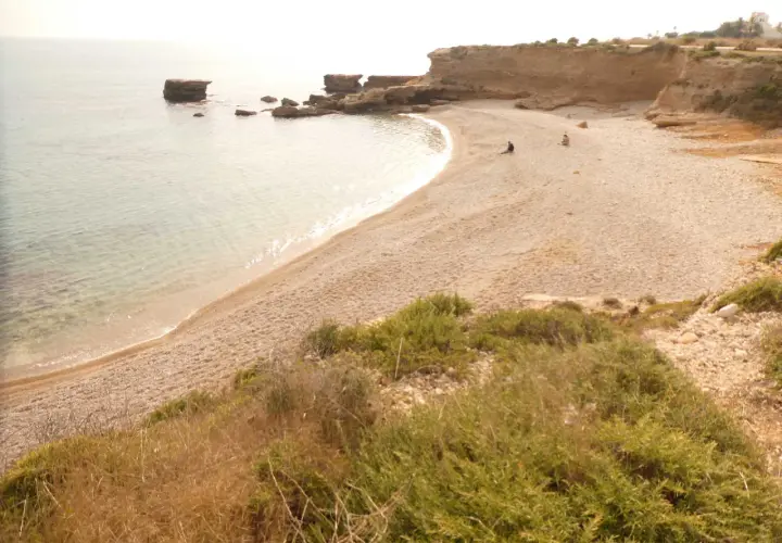 Una tranquila cala foradada con aguas cristalinas y rocas emergiendo del mar. Dos personas disfrutan de la soledad en la playa de guijarros.