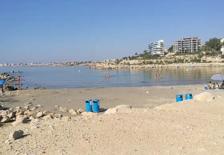 Día soleado en la cala Morro Blanc. Gente disfrutando del mar y la playa, con edificios modernos al fondo.