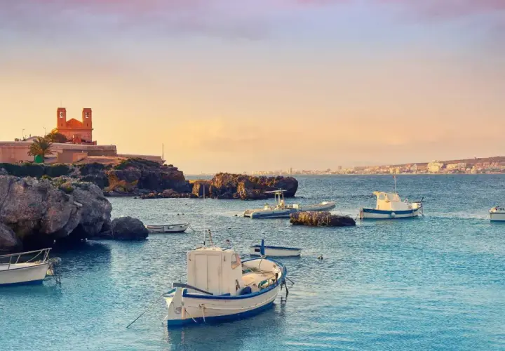 Barcas tranquilas en el puerto Santa Pola, con edificios históricos y la costa al fondo bajo un cielo anaranjado.