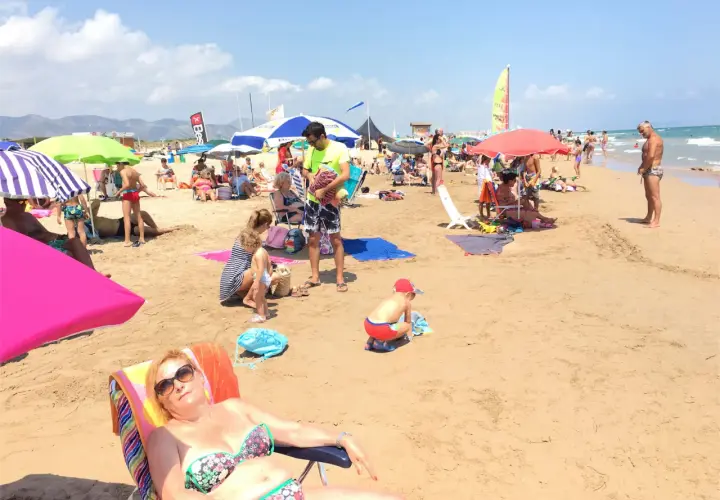 Día soleado en la playa de l'Ahuir, con gente disfrutando bajo sombrillas y el mar al fondo.