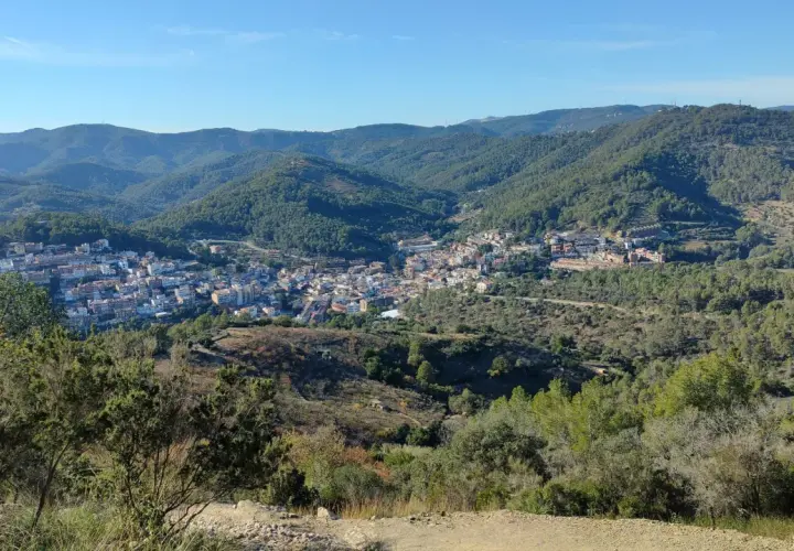 Vista panorámica de un pueblo en un valle rodeado de montañas boscosas. Se distingue la ermita de Sant Ramon.