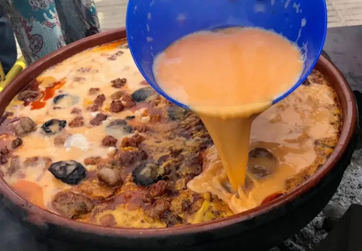 Un líquido naranja se vierte de un recipiente azul a una paella con carne y marisco.