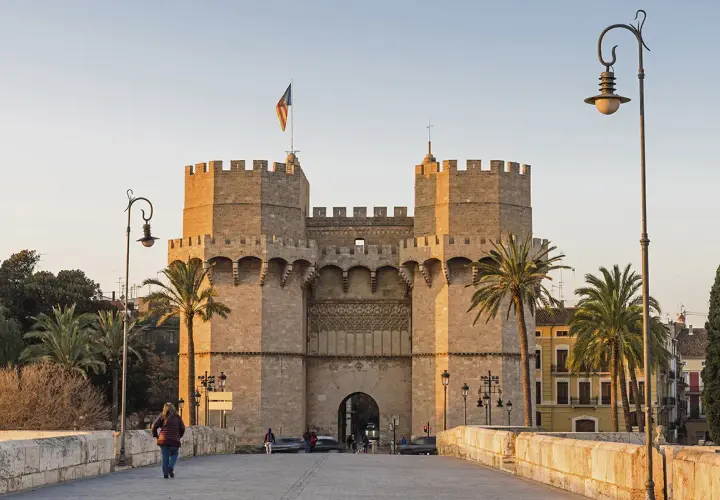 Las **Torres de Serranos**, imponentes puertas de la antigua muralla de Valencia, se alzan majestuosas bajo un cielo despejado. Palmeras y farolas flanquean el camino.