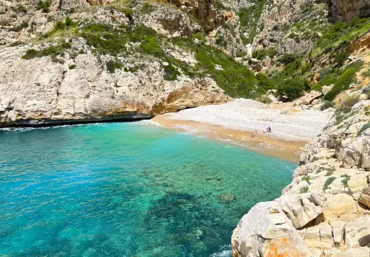 Aguas turquesas bañan la cala testos, una playa de guijarros rodeada de acantilados rocosos y vegetación.