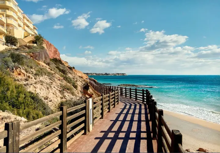 Paseo de madera con vistas a las mejores playas de Cabo Roig. Mar turquesa, arena dorada y un edificio amarillo en la ladera.