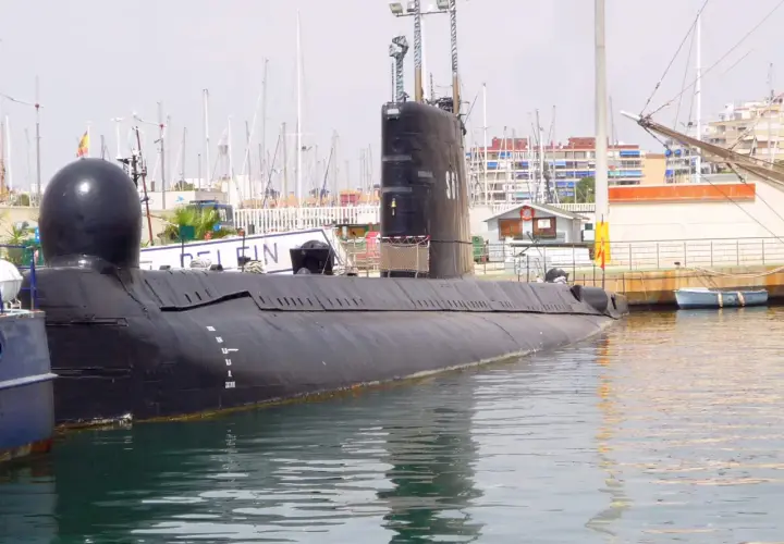 Un submarino negro, el Delfín, descansa en el puerto de Torrevieja, con mástiles de veleros y edificios al fondo.