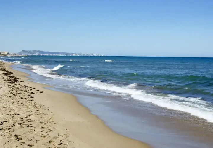 La playa de Xeraco, con arena dorada y el mar azul, se extiende hacia un horizonte con edificios y montañas.
