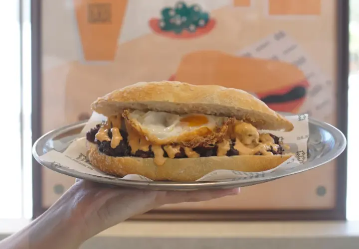 Un delicioso almuerzo valenciano: un bocadillo con carne, huevo frito y salsa cremosa, servido en plato metálico.