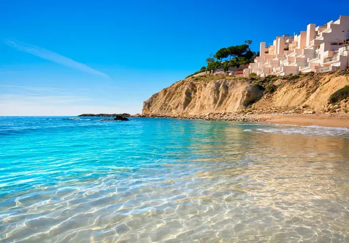 Aguas cristalinas en las calas de Alicante, con una urbanización blanca en la cima del acantilado y el mar azul intenso.