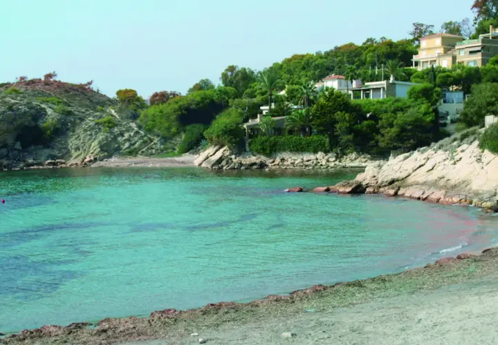 Aguas turquesas bañan la cala d'enmig, con casas blancas y verdes colinas de fondo.