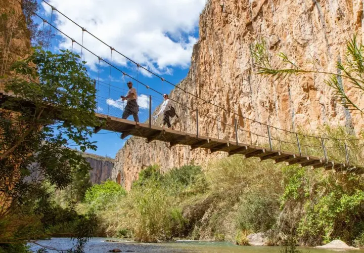 Dos excursionistas cruzan un puente colgante sobre un río, explorando las rutas de senderismo de Valencia.