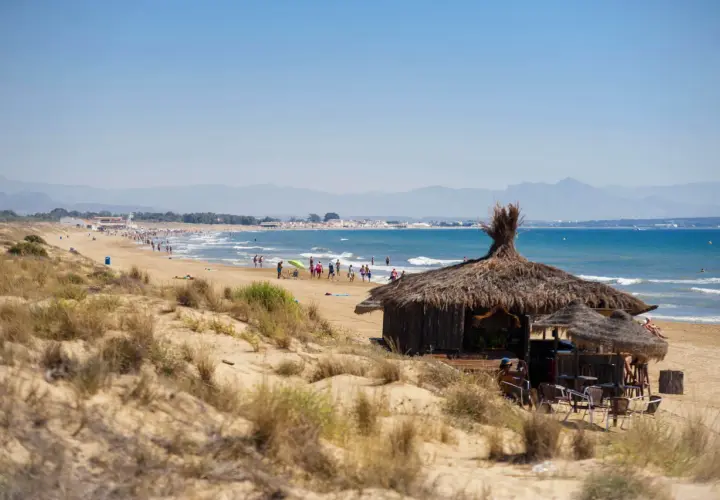 Chiringuito de paja en la playa del Rebollo, con gente disfrutando del sol y el mar azul. Montañas al fondo.
