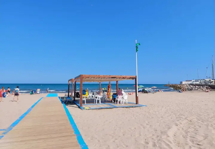 Pasarela de madera azul en la playa de Massamagrell, con chiringuito y gente disfrutando del sol y el mar.