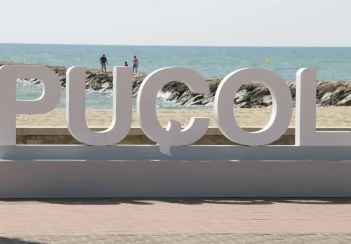 Letras gigantes "PUZOL" en la playa, con el mar azul y personas al fondo.