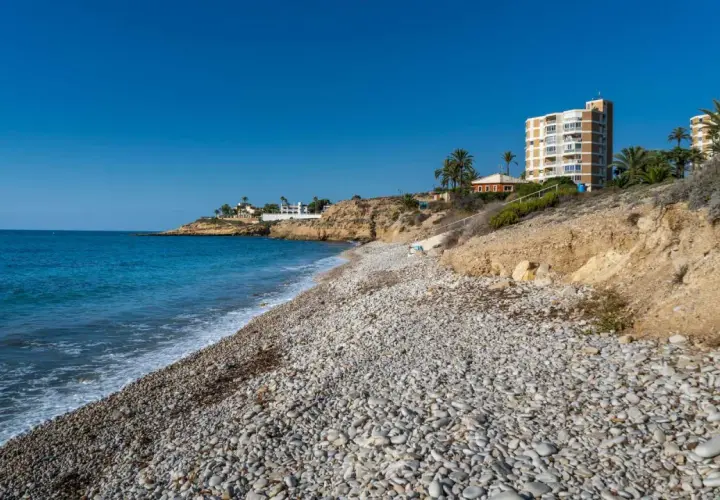 La cala Amerador, con su playa de guijarros y aguas turquesas, se extiende bajo un cielo azul. Edificios modernos y vegetación mediterránea bordean la costa.