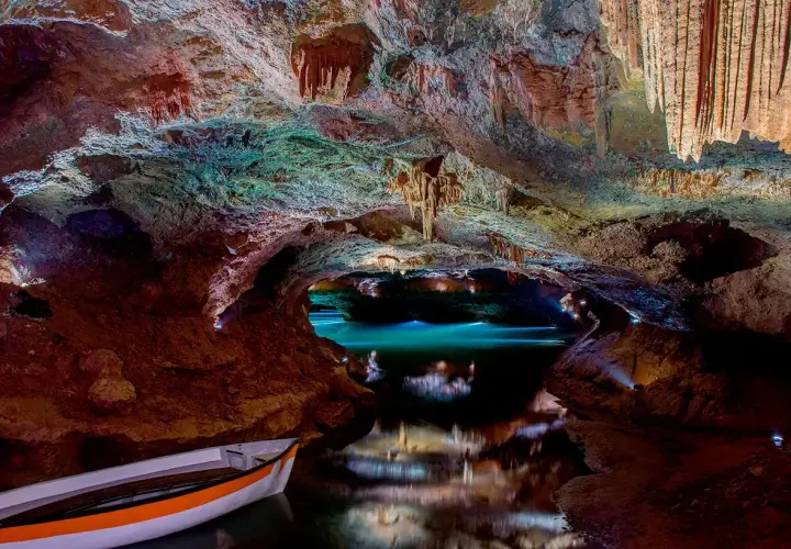 Interior de cueva con estalactitas, agua turquesa y una barca. Consulta las cuevas de Vall d'Uixó horarios y precios para tu visita.