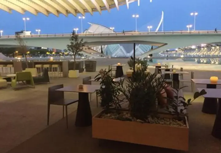 Mesas y sillas en la terraza de restaurantes con vistas a un puente moderno y un cielo azul crepuscular.