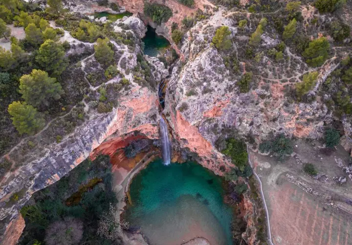 Cascada cayendo en una poza turquesa, rodeada de rocas rojizas y vegetación. Un rincón secreto, como la cueva turche, esperando ser descubierto.