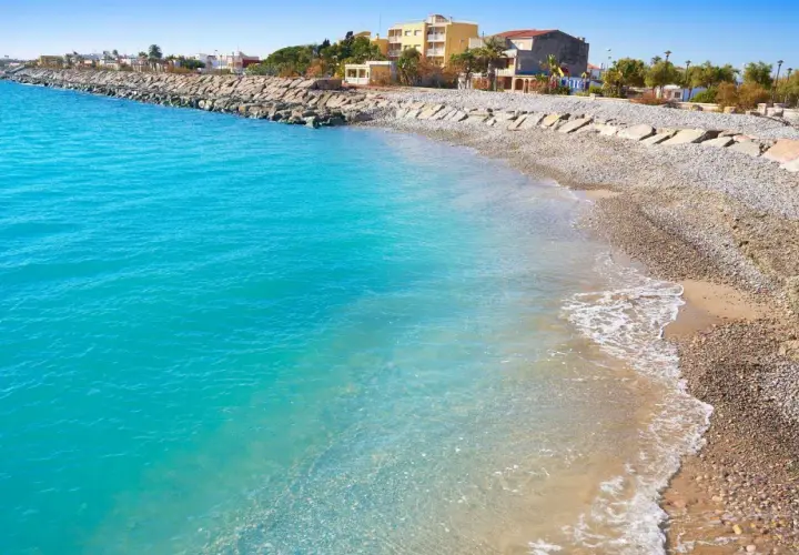 Aguas turquesas bañan la playa almenara, con un rompeolas de rocas y edificios al fondo.