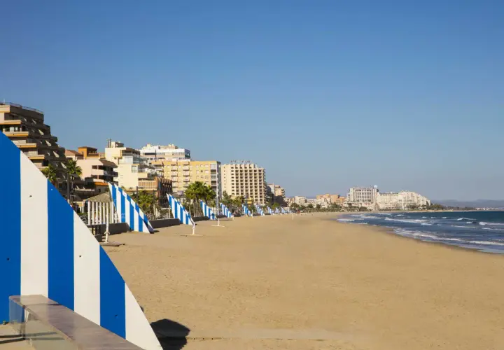 Amplia playa del Moro con edificios turísticos al fondo y sombrillas a rayas azules y blancas alineadas en la arena dorada.