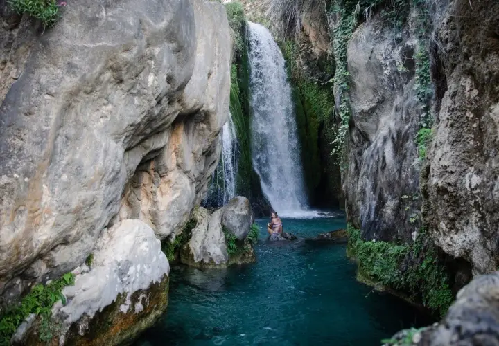 Una mujer se baña en las **fonts de Algar**, rodeada de rocas y vegetación, bajo una cascada de agua cristalina.