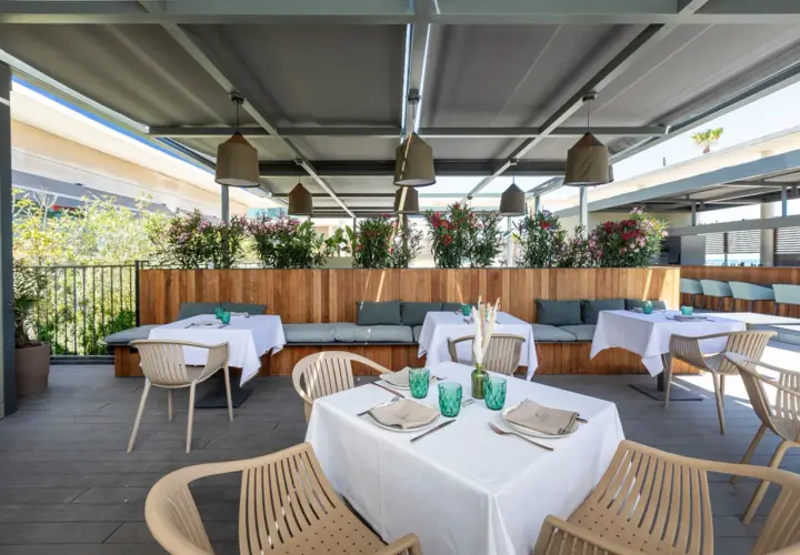 Terraza de la colonia de carmela con mesas puestas, asientos de madera y plantas floridas.
