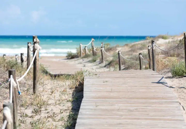 Pasarela de madera que conduce a la playa Raco de la Mar. El mar azul y las dunas de arena crean un paisaje sereno.