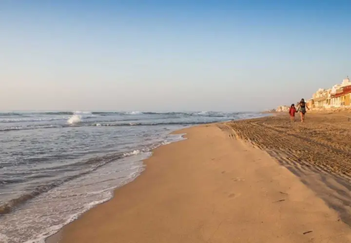 Dos personas pasean por la **playa Babilonia** al atardecer, con el mar y las casas de fondo.