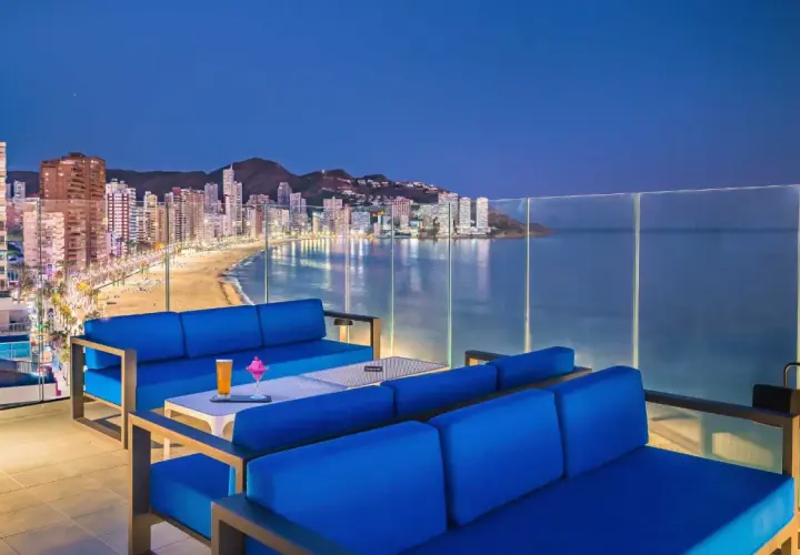 Sofás azules en terraza con vistas a la playa y la ciudad iluminada de Barceló Benidorm Beach al anochecer.
