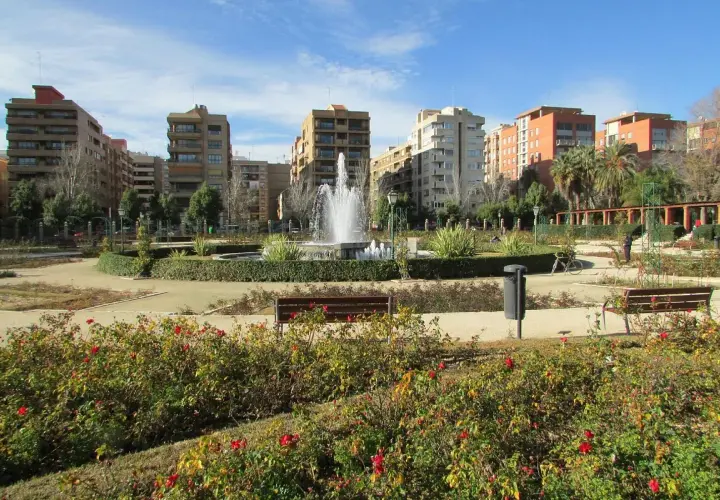 Parque urbano con fuente central y edificios residenciales al fondo. Los viveros de Valencia lucen sus rosales en flor.
