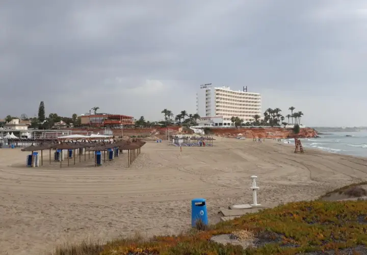 Playa con sombrillas y tumbonas preparadas. Al fondo, un hotel blanco y edificios costeros. El mar se ve tranquilo bajo un cielo nublado. Un día perfecto para disfrutar de la cala del bosque.