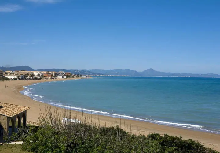 Amplia vista de la playa de Les Bovetes con arena dorada, mar azul y montañas al fondo. Un día soleado perfecto para disfrutar.