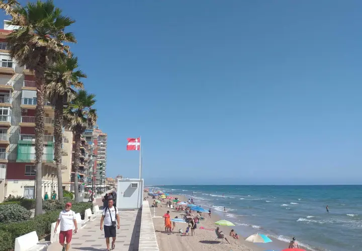 Gente disfrutando del sol y el mar en la playa Mareny de Barraquetes, con palmeras y edificios de apartamentos.
