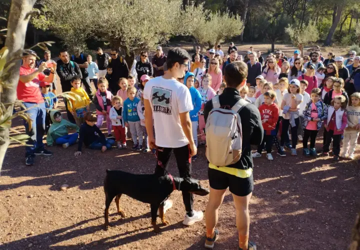 Un grupo de personas, incluyendo niños, se reúne en un sendero boscoso. Un hombre con una camiseta roja toma una foto con su móvil. Un perro Dóberman negro está atado a un joven. Quizás están buscando al **dragón de la Calderona**.