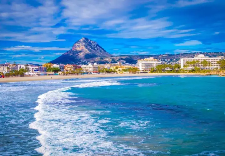 Olas turquesas rompen en la arena dorada de la playa del Arenal Javea, con edificios y el imponente Montgó al fondo.