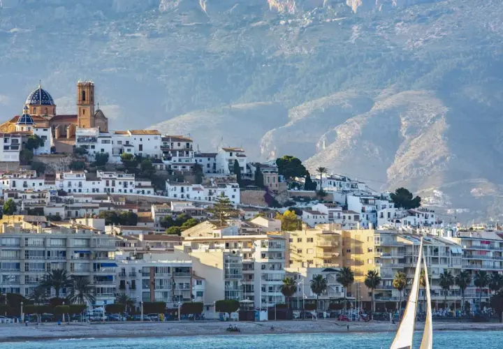 Vista del **castell de l'Olla** coronando la colina, con sus casas blancas y la iglesia de cúpula azul. Un velero navega frente a la costa.