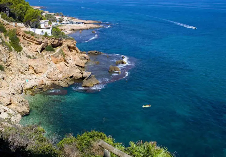 Vista de la cala Les Rotes con aguas cristalinas, acantilados rocosos y un kayak amarillo en el mar azul.