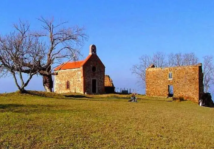 La ermita de Santa Catalina, una pequeña iglesia de piedra con tejado naranja, se alza junto a unas ruinas bajo un cielo azul. Un árbol desnudo y un paisaje verde completan la escena.