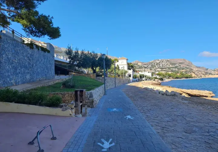 Paseo marítimo con siluetas de personas en la cala del Soio. El mar azul y las montañas de fondo crean un paisaje idílico.