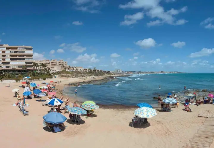 Gente disfrutando del sol y el mar en la playa Cabo Cervera, con sombrillas de colores y edificios al fondo.