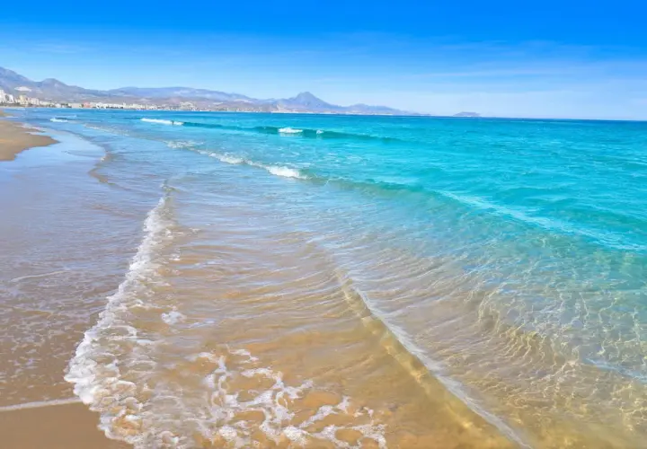 Aguas turquesas y arena dorada en las calas de San Juan, Alicante. El mar Mediterráneo acaricia la costa, con montañas al fondo.