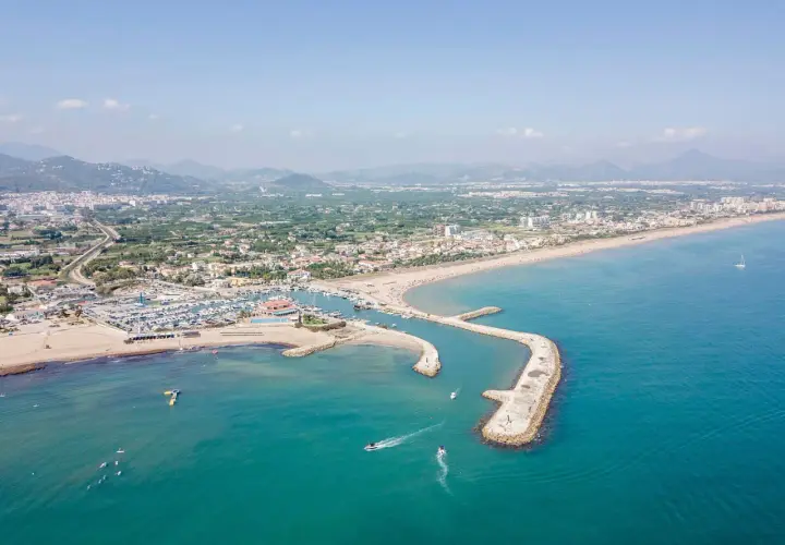 Vista aérea del puerto de Oliva con barcos navegando y la playa llena de gente.