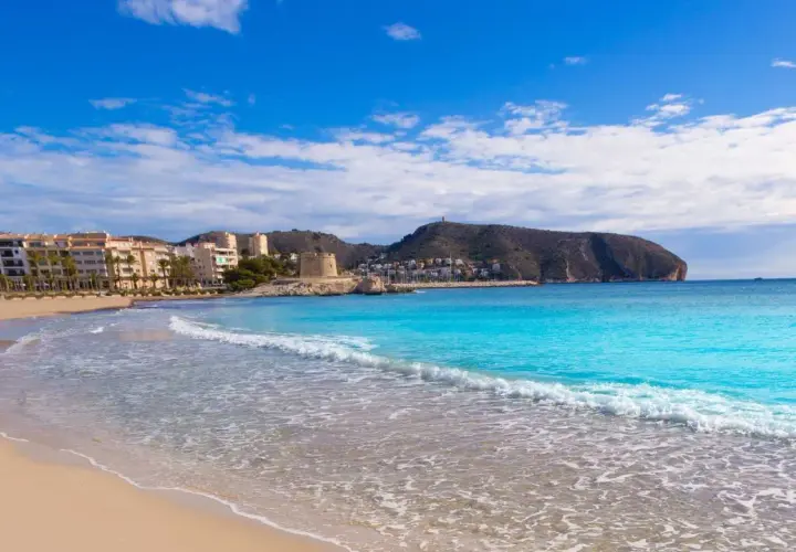 La playa de l'Ampolla en Moraira, con su arena dorada y aguas turquesas, invita a un día de sol y mar. Al fondo, edificios y una torre defensiva se integran en el paisaje.