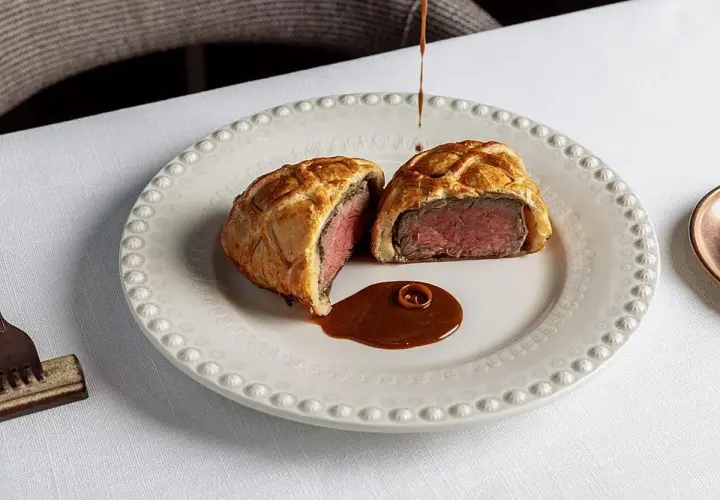 Un Wellington de ternera jugoso, cortado por la mitad, servido con salsa en un plato elegante. Una delicia para probar en los restaurantes de Valencia.