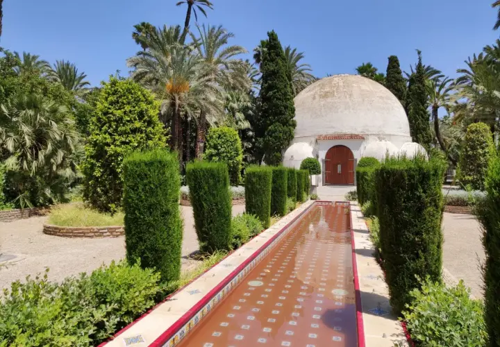 Un estanque de agua rojiza y un edificio abovedado en el parque el palmeral, rodeado de palmeras y vegetación exuberante.