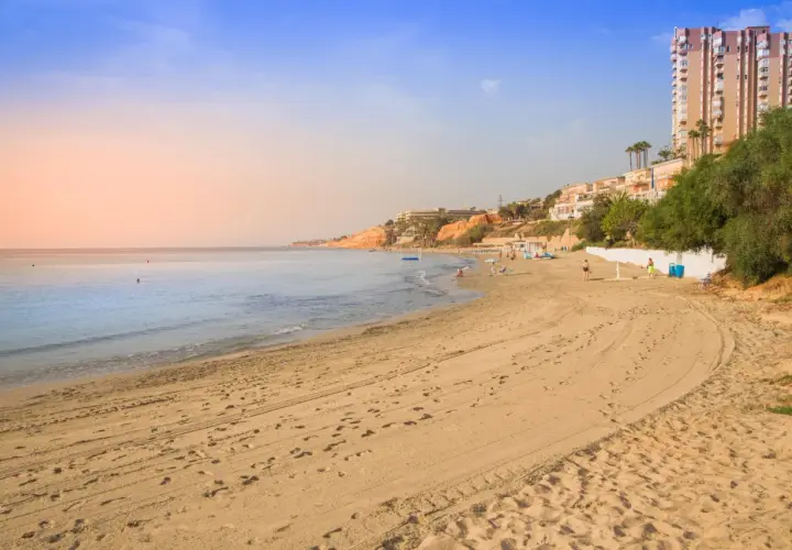 Playa con arena dorada y el mar azul. Al fondo, un barranco rubio con edificios y vegetación.