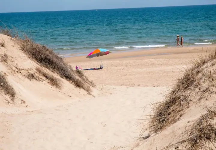 Dunas de arena y mar azul en el mareny de Sant Llorenç. Una sombrilla colorida y dos personas pasean por la orilla.