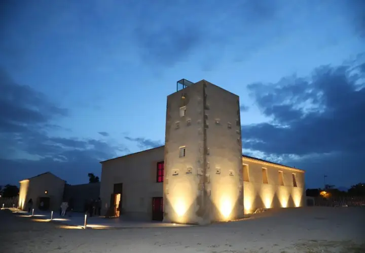 La torre Ansaldo, iluminada al anochecer, se alza junto a edificios bajos bajo un cielo azul oscuro con nubes.