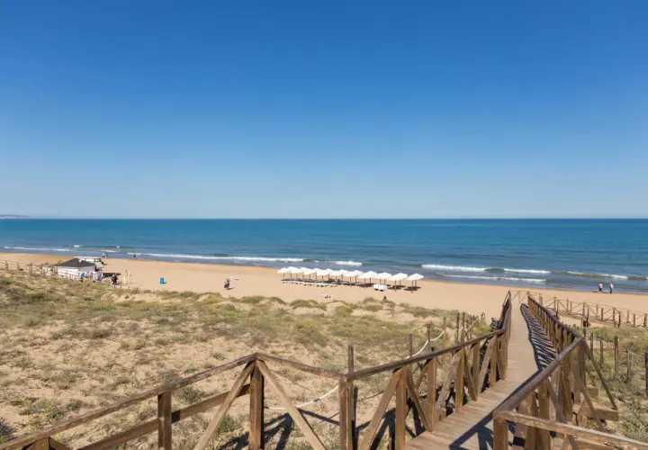 Pasarela de madera hacia la playa Moncayo, con arena dorada, mar azul y sombrillas blancas alineadas.